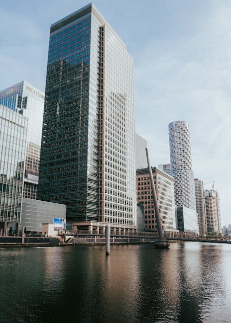 The image depicts a modern cityscape featuring a collection of tall office buildings with glass and steel facades, situated along a river. The main building in the foreground is a rectangular skyscrap