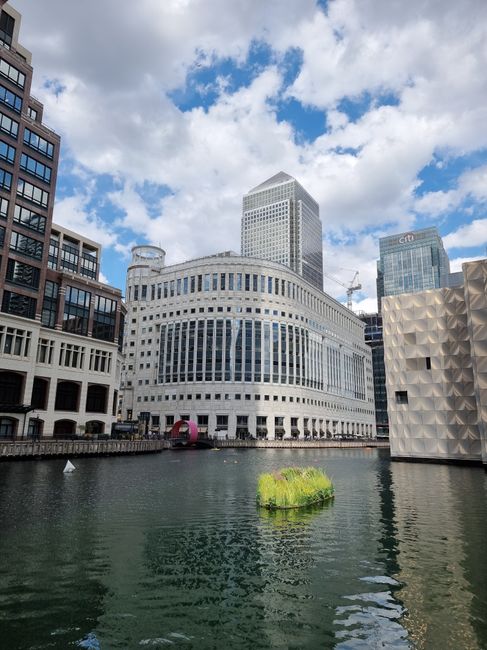 The image depicts a modern cityscape featuring a collection of tall office buildings with glass and steel facades, situated along a river. The main building in the foreground is a rectangular skyscrap